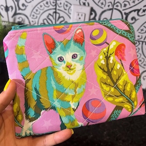May include: Pink quilted zippered pouch featuring a colorful cat illustration with blue, green, and yellow stripes. The design includes stars and lollipop-like shapes. The pouch has a teal zipper and is held in a hand.