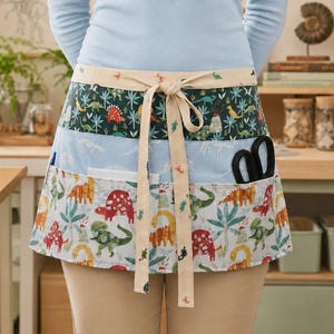 May include: A waist apron featuring three horizontal bands with dinosaur and tropical plant patterns in shades of green, blue, red, and yellow. The apron has a cream-colored tie at the waist and two front pockets containing a pair of black scissors.