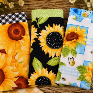 May include: Three rectangular fabric cases for eyeglasses or small items. The left case features yellow sunflowers with a black and white gingham top. The center case has yellow sunflowers on black with a green polka dot top. The right case displays sunflowers, butterflies, and bees on white.