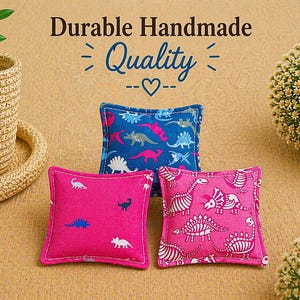 May include: Three small square fabric cushions featuring dinosaur patterns. One cushion is blue with multi-colored dinosaurs, while two are pink with white dinosaur silhouettes and skeletal prints. Text above reads Durable Handmade Quality.