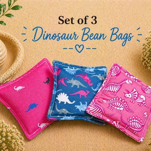 May include: A set of three square fabric bean bags featuring dinosaur patterns. One bag is bright pink with small white and blue dinosaur silhouettes, one is blue with multi-colored dinosaur shapes, and one is pink with white dinosaur skeletons.