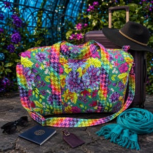 May include: A quilted duffel bag with a vibrant floral pattern in shades of purple, pink, and green, featuring a colorful houndstooth strap. The set includes a black suitcase, a black hat, a dark blue passport holder, sunglasses, and a teal scarf.