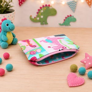 May include: A rectangular fabric zipper pouch featuring a colorful dinosaur print with pink, blue, and green patterns. The interior lining is blue with white polka dots. The pouch is positioned on a wooden surface near a small plush dinosaur toy.