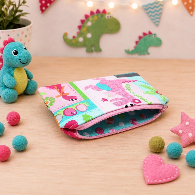 May include: A rectangular fabric zipper pouch featuring a colorful dinosaur print with pink, blue, and green patterns. The interior lining is blue with white polka dots. The pouch is positioned on a wooden surface near a small plush dinosaur toy.