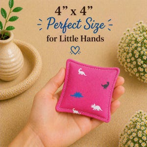 May include: A square pink fabric bean bag featuring a repeating pattern of small white and blue dinosaur silhouettes. The item measures 4 inches by 4 inches as indicated by the text printed above it, which also reads Perfect Size for Little Hands.