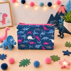 May include: A rectangular zippered pouch featuring a dark blue fabric base with a repeating pattern of various dinosaurs in shades of pink, light blue, and white. The pouch has a bright pink zipper closure along the top edge.