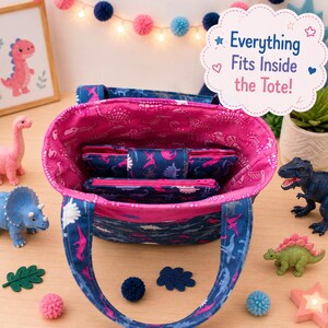 May include: A fabric tote bag with a dark blue exterior featuring a dinosaur pattern and a bright pink interior lining with a dinosaur skeleton print. The bag contains a matching zippered pouch and a folded wallet, with text reading Everything Fits Inside the Tote!