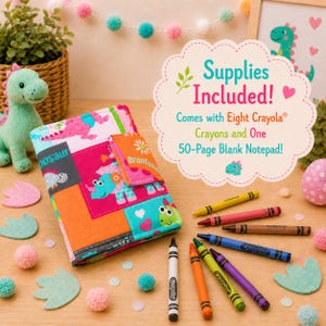 May include: A fabric notebook cover featuring a colorful dinosaur print in shades of pink, orange, and teal. The cover includes a flap closure and is accompanied by eight Crayola crayons. Text on the included sign reads: Supplies Included! Comes with Eight Crayola Crayons and One 50-Page Blank Notepad!