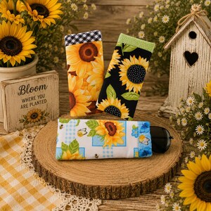 May include: Three rectangular fabric cases featuring sunflower patterns. One case has a yellow sunflower print with a black and white gingham border, another has a black background with sunflowers, and the third is white with sunflowers and bees.