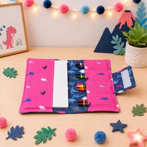 May include: A pink fabric crayon roll featuring a dinosaur pattern, containing a white rectangular notepad and six colorful crayons in individual slots. The roll has a blue interior lining with a dinosaur print and a hook-and-loop closure tab.