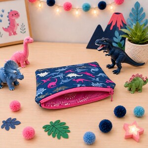 May include: A rectangular fabric zipper pouch featuring a dark blue exterior with a repeating pattern of pink, light blue, and white dinosaurs. The interior lining is bright pink and decorated with a white dinosaur print. The zipper is pink.