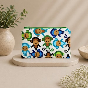 May include: A white zippered pouch with a repeating pattern of smiling cartoon monkeys in various colors. The zipper is green, and the pouch is rectangular. The monkeys have brown, blue, and green faces.
