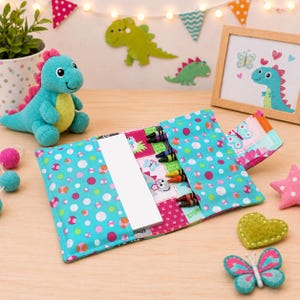 May include: A fabric roll-up case featuring a turquoise background with multicolored polka dots, containing a white notepad and several crayons. The case has interior pockets for organizing art supplies and a flap closure with a butterfly print pattern.