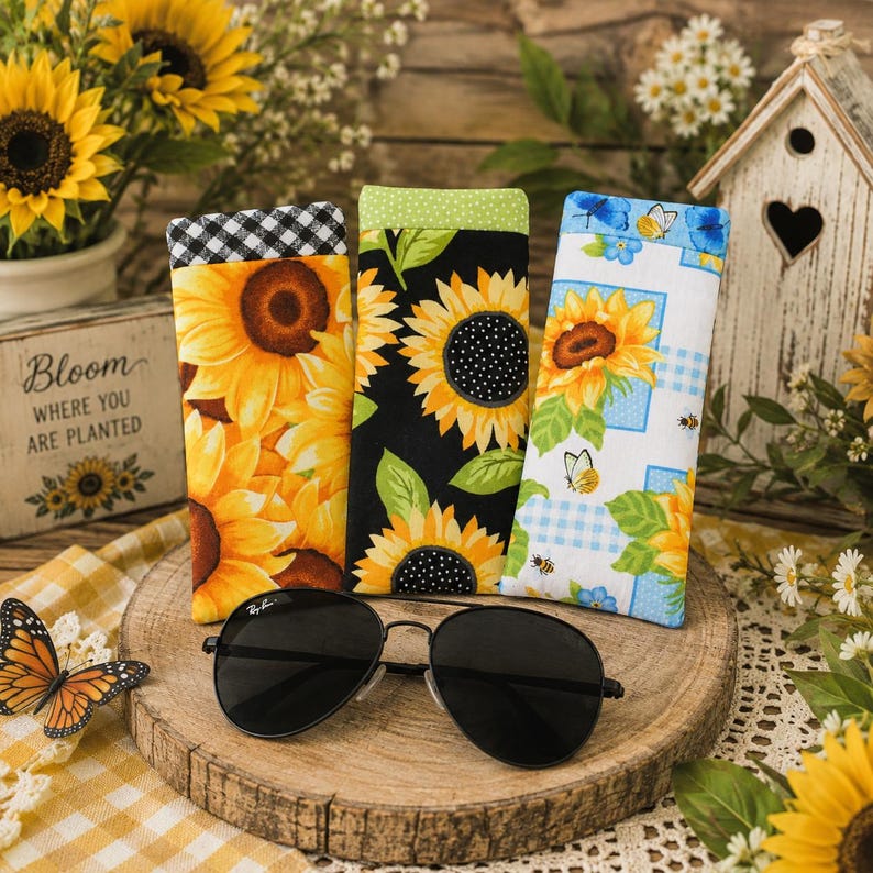 May include: Three rectangular fabric eyeglass cases featuring sunflower patterns. One case has a black and white gingham top, one has a green polka dot top, and one has a blue and white checkered background with butterflies and bees.