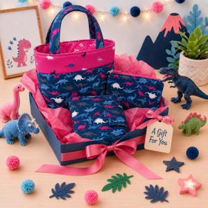 May include: A fabric gift set featuring a dark blue tote bag, a zippered pouch, and a folded wallet, all decorated with a colorful dinosaur pattern. The items are arranged in a dark blue box with pink tissue paper and a pink ribbon with a tag reading A Gift For You.