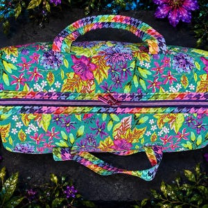 May include: A rectangular fabric bag featuring a vibrant floral pattern in shades of teal, magenta, purple, and yellow. The bag includes a top zipper closure and two handles, both decorated with a multi-colored houndstooth pattern in rainbow hues.