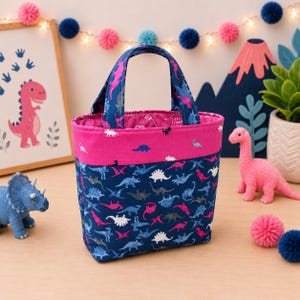 May include: A fabric tote bag featuring a dark blue background with a repeating pattern of small dinosaurs in pink, white, and light blue. The bag has a solid bright pink horizontal band near the top and two matching fabric handles.