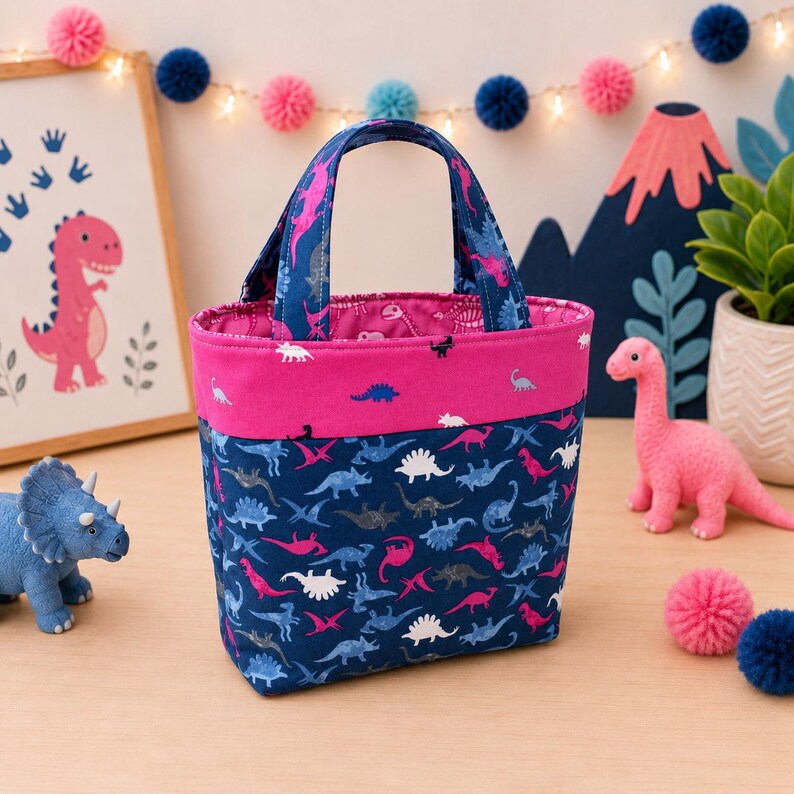 May include: A fabric tote bag featuring a dark blue background with a repeating pattern of small dinosaurs in pink, white, and light blue. The bag has a solid bright pink horizontal band near the top and two matching fabric handles.