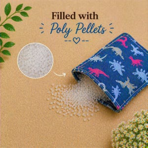 May include: A blue fabric pouch featuring a repeating pattern of pink and light blue dinosaurs. The pouch is partially open, spilling small white poly pellets onto the surface. Text above reads: Filled with Poly Pellets with a heart icon.