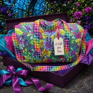 May include: A quilted duffel bag featuring a vibrant floral pattern in shades of teal, purple, and green with a multicolored houndstooth strap. A rectangular gift tag attached to the handle displays the text A Gift for You in black font.