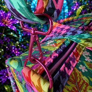 May include: A quilted fabric bag featuring a vibrant floral pattern in shades of green, pink, and purple, accented by a colorful houndstooth print on the strap. The bag includes a metallic pink swivel clasp and D-ring hardware with a zipper closure.