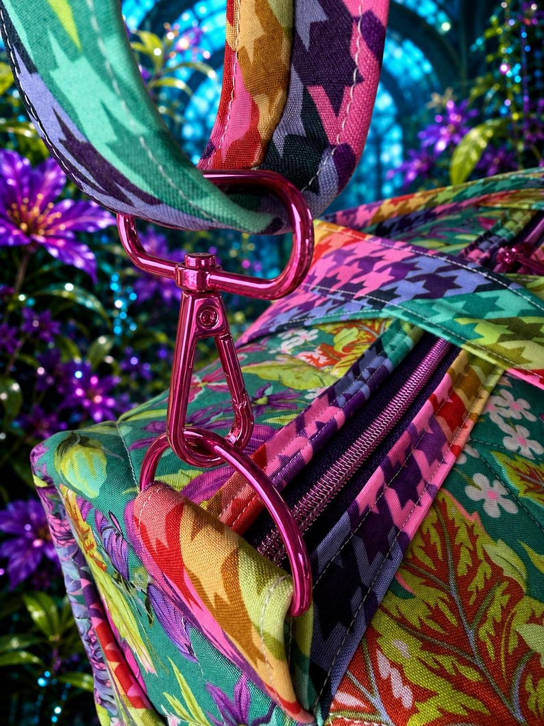 May include: A quilted fabric bag featuring a vibrant floral pattern in shades of green, pink, and purple, accented by a colorful houndstooth print on the strap. The bag includes a metallic pink swivel clasp and D-ring hardware with a zipper closure.