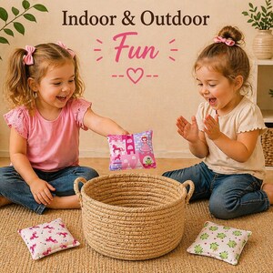 May include: A round woven rope basket with two handles, accompanied by three small square fabric bean bags featuring patterns of unicorns, castles, and green frogs. The text Indoor & Outdoor Fun is displayed above the items.