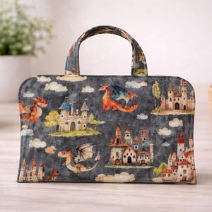 May include: A rectangular tote bag with a handle, featuring a fantasy design. The bag is dark gray with illustrations of castles, dragons, and clouds. The dragons are in shades of orange, red, and blue. The castles have towers and flags.