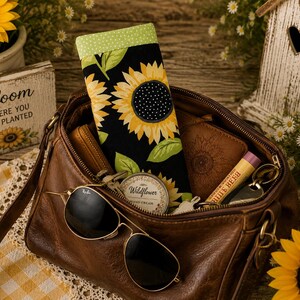 May include: A brown leather handbag containing a fabric eyeglass case with a sunflower pattern on a black background, a round Wildflower hand cream tin, a tube of Burt's Bees lip balm, a brown wallet, and black aviator sunglasses resting on top.
