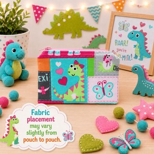 May include: A rectangular fabric pouch featuring a colorful patchwork design with a green dinosaur, pink hearts, and a butterfly. The pouch has a zipper closure and displays various patterns including a small dinosaur hatching from an egg.