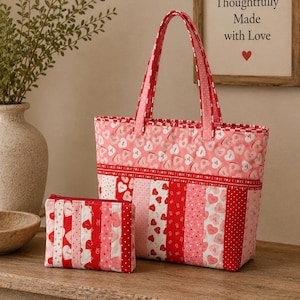 May include: A quilted tote bag and matching zippered pouch. The bag and pouch feature a patchwork design with red, pink, and white fabrics. The fabrics are patterned with hearts and the words "I love you". The tote bag has pink handles and a red trim. The framed text reads "Thoughtfully Made with Love".