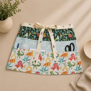 May include: A colorful half apron with a dinosaur theme. The apron features three horizontal sections with different patterns: a dark green section with dinosaurs and trees, a light blue section with dinosaur silhouettes, and a white section with various dinosaurs and plants. A cream-colored tie and pockets complete the design.