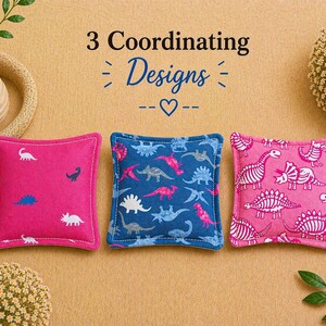 May include: Three square fabric sachets featuring dinosaur patterns. The left sachet is hot pink with small white and blue dinosaurs. The center sachet is navy blue with multi-colored dinosaurs. The right sachet is pink with white dinosaur skeletons.
