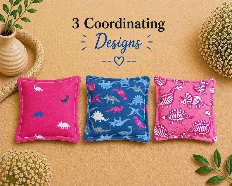 May include: Three square fabric sachets featuring dinosaur patterns. The left sachet is hot pink with small white and blue dinosaurs. The center sachet is navy blue with multi-colored dinosaurs. The right sachet is pink with white dinosaur skeletons.