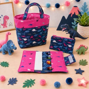 May include: A fabric tote bag, a zippered pouch, and a folding crayon roll, all featuring a dark blue and pink dinosaur pattern. The set includes a white notepad and several colorful crayons tucked into the slots of the open crayon roll.