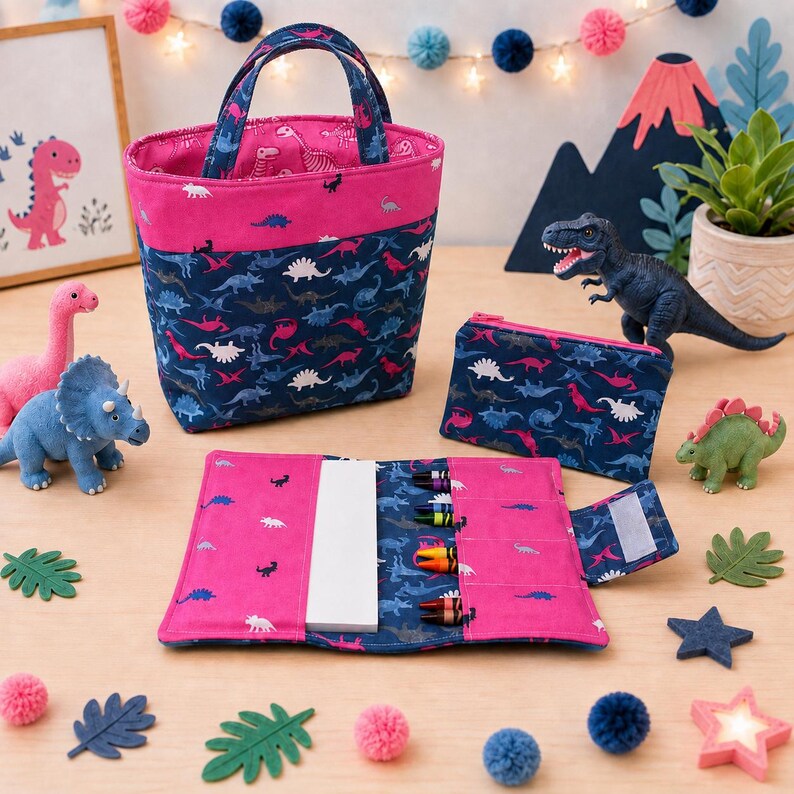 May include: A fabric tote bag, a zippered pouch, and a folding crayon roll, all featuring a dark blue and pink dinosaur pattern. The set includes a white notepad and several colorful crayons tucked into the slots of the open crayon roll.