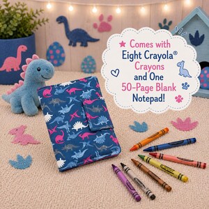 May include: A blue fabric notebook cover featuring a dinosaur pattern, accompanied by eight Crayola crayons. A white scalloped sign reads: Comes with Eight Crayola Crayons and One 50-Page Blank Notepad! The set includes various colored wax crayons.