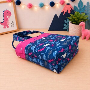 May include: A fabric tote bag featuring a dark blue background with a repeating pattern of dinosaurs in shades of pink, light blue, and white. The bag includes a solid bright pink fabric band across the top and matching pink fabric handles.