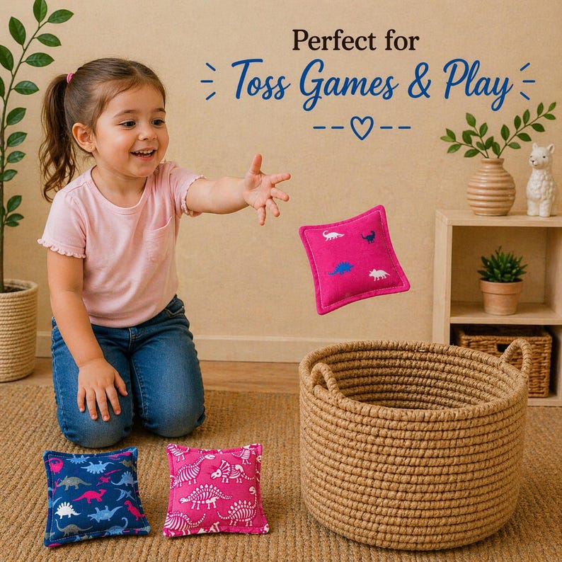 May include: A set of square fabric bean bags featuring dinosaur patterns in pink and blue, accompanied by a round woven rope basket. The text Perfect for Toss Games & Play is visible above the items, along with a small heart icon.