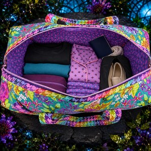 May include: A quilted duffel bag with a vibrant floral pattern in shades of teal, pink, and yellow, featuring a purple polka dot interior. The bag is packed with folded clothing, including a black shirt, a teal shirt, and purple patterned pajamas.