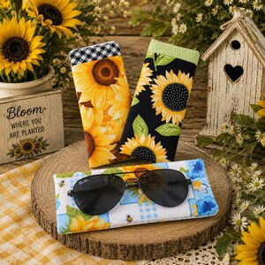 May include: Three rectangular fabric pouches featuring sunflower patterns. Two pouches stand upright, one with a yellow sunflower print and black-and-white gingham trim, the other with a black background and sunflowers. A third pouch lies flat with blue accents.