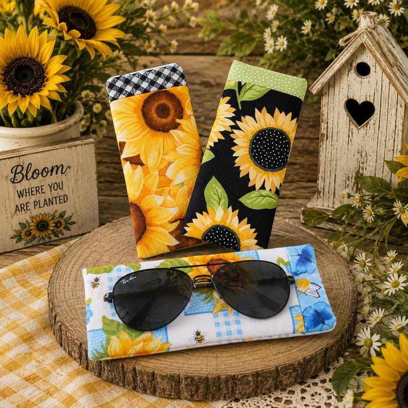 May include: Three rectangular fabric pouches featuring sunflower patterns. Two pouches stand upright, one with a yellow sunflower print and black-and-white gingham trim, the other with a black background and sunflowers. A third pouch lies flat with blue accents.