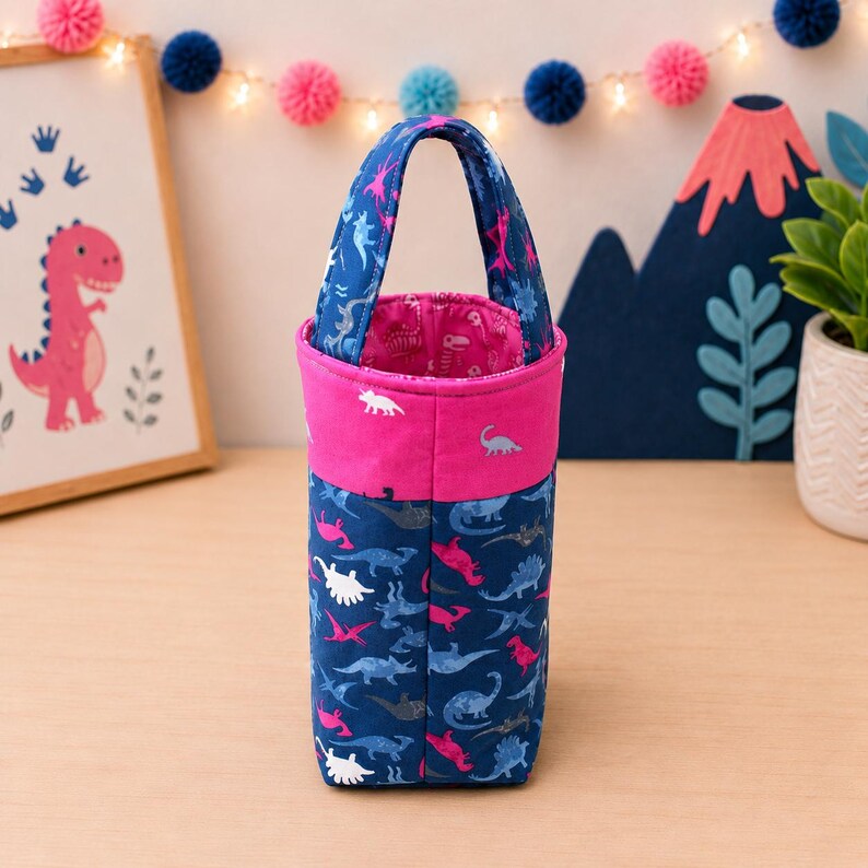 May include: A cylindrical fabric bag with a handle, featuring a dark blue exterior with a repeating pattern of various dinosaurs in white, light blue, and pink. The top section is a solid pink band, and the interior lining is pink with dinosaurs.