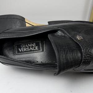 May include: Close-up of a black leather shoe with the "Gianni Versace" label visible inside. The shoe features a textured surface and a decorative metal emblem. The sole is visible in the background.