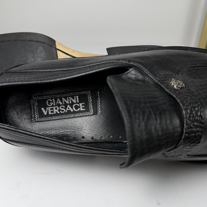 May include: Close-up of a black leather shoe with the "Gianni Versace" label visible inside. The shoe features a textured surface and a decorative metal emblem. The sole is visible in the background.