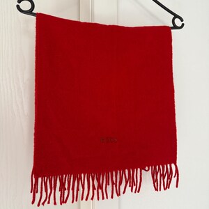 May include: A vibrant red scarf with fringe detail, hanging on a black hanger. The scarf has the word "BOSS" embroidered on it. This accessory is perfect for adding a pop of color to any outfit.