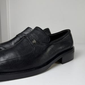 May include: A pair of black leather loafers. The shoes have a textured appearance and a small decorative emblem on the strap. The soles and heels are also black. The shoes are displayed on a white surface.