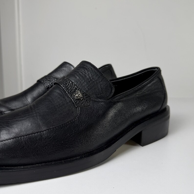 May include: A pair of black leather loafers. The shoes have a textured appearance and a small decorative emblem on the strap. The soles and heels are also black. The shoes are displayed on a white surface.