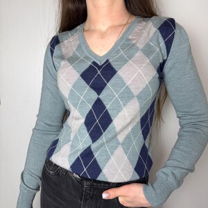 May include: A long-sleeved V-neck sweater with a traditional argyle design. The sweater showcases a pattern of blue, grey, and navy diamonds, edged with white lines. The sleeves and neckline are a solid blue, providing a contrast to the patterned body.