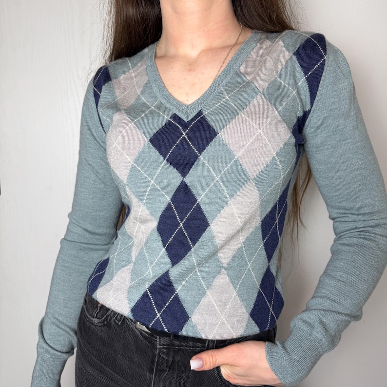 May include: A long-sleeved V-neck sweater with a traditional argyle design. The sweater showcases a pattern of blue, grey, and navy diamonds, edged with white lines. The sleeves and neckline are a solid blue, providing a contrast to the patterned body.